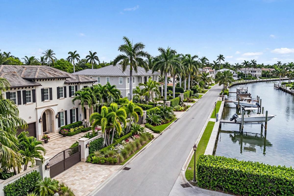 A photorealistic view of an upscale residential street in Old Naples, Port Royal, Aqualane Shores, Naples, Florida, lined with coastal estate homes and mature landscaping on a clear day.